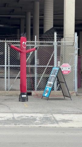 The scene is an urban outdoor environment, with a red inflatable tube man standing to the left of a sign that reads "Lave Auto A la Main $29.99". The tube man has a smiley face on its body. A silver car passes in front of the tube man and sign, moving from right to left. In the background, a chain-link fence runs horizontally across the view, with concrete pillars and parts of a building visible behind it. There is also a surveillance warning sign attached to the fence. The ground is paved.