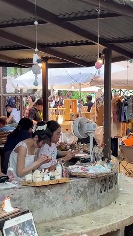 The video captures an outdoor setting under a corrugated metal roof, suggesting a market or craft fair. In the foreground, two young women are seated at a circular concrete counter, actively engaged in painting small ceramic figures. The woman on the left, wearing a white dress with thin straps, has her hair pulled back with a white scrunchie. She is holding a small brush and appears to be applying paint to a white figure. To her right, another woman, also wearing a white top, is focused on her own ceramic figure, holding a small brush and paint. The counter is cluttered with various art supplies, including paint pots, brushes, and more unfinished ceramic figures. A white oscillating fan is positioned behind the women on the counter, likely for ventilation.

Behind them, other individuals are visible, browsing or working at stalls. To the right, a rack displays clothing items, including shirts and dresses, suggesting it's a retail area. In the background, white umbrellas are open, providing shade, and there are glimpses of greenery, indicating an outdoor environment. The overall atmosphere appears relaxed and creative, with people enjoying an outdoor marketplace activity.