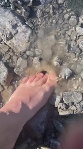 A close-up, top-down shot shows a bare foot stepping into shallow, murky water. The foot is light-skinned, and the toes are visible. The water is surrounded by numerous gray and white rocks of varying sizes. Ripples are visible in the water as the foot makes contact. The overall scene suggests an outdoor, natural environment.