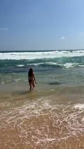 The video shows a person wearing a dark-colored bikini standing on a sandy beach. The person, who appears to be female, is facing the ocean and is standing in the shallow water where the waves meet the shore. The water is clear enough to see the sandy bottom. The waves are moderate in size, with white foam crests. The sky is clear and blue, and the sun is shining brightly. The beach appears to be relatively deserted. The woman then begins to walk further down the beach.