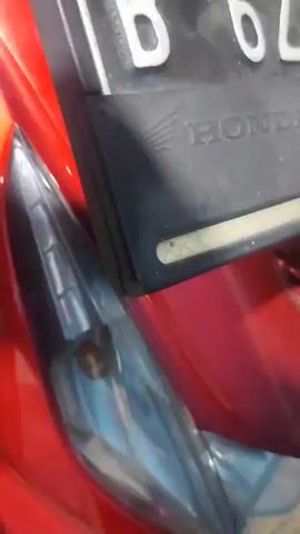 The media shows a close-up, medium, and wide shot of a bright red motorcycle. The motorcycle is parked indoors, possibly in a garage or storage area, as evidenced by the concrete floor and some visible pipes and a broom in the background. The motorcycle has black accents on its seat and body, and the license plate is clearly visible, showing the text "B 6289 CTT" and "HONDA". The lighting suggests it is daytime, but the interior setting makes it hard to determine the exact time of day or weather. The motorcycle appears to be stationary, with no activity or event taking place other than its being photographed. The focus is on the details of the motorcycle's design, including its headlights and body panels, highlighting its glossy red finish.