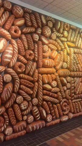 A video displays a wall covered in various baked goods, appearing to be made of ceramic or plaster. The items are arranged in a dense, decorative pattern and include loaves of bread, rolls, and pretzels in different shapes and sizes. The baked goods are predominantly in shades of brown and tan, mimicking the appearance of freshly baked bread. The lighting suggests an indoor setting, possibly a bakery or a decorative installation. The camera pans across the wall, offering a close-up view of the textured details of each item. There are no people visible in the video, and the focus remains solely on the bread-themed wall decoration.