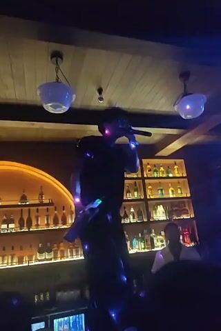 A person dressed in dark clothing, including a t-shirt and pants, stands on top of a bar counter. They are holding a microphone to their mouth and appear to be singing or speaking into it. The person is illuminated by various colored lights, which appear as blue and purple spots on their clothing and body. Behind the person is a well-stocked bar with shelves of bottles, illuminated by warm lighting. Two hanging light fixtures with white glass shades are visible on the wooden ceiling. Another person in a white shirt is standing behind the bar counter, facing the person on top. The scene appears to be a dimly lit bar or nightclub environment.