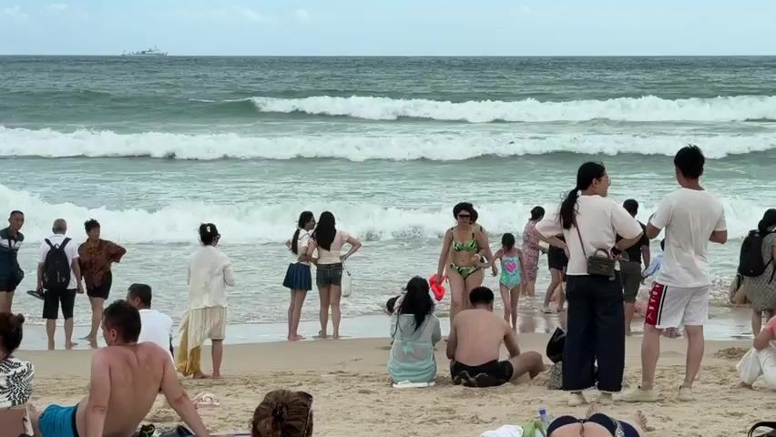 The video depicts a crowded beach scene with numerous people scattered across the sand and standing near the water. The ocean features moderate waves, and a distant ship is visible on the horizon. Most individuals are dressed in casual beach attire, including swimwear and light clothing. Some are sitting or lying on the sand, while others are standing and interacting. Several groups are conversing, and some appear to be taking photographs. A few people are wading in the shallow water. The sky is overcast, suggesting a cloudy day. The camera perspective changes slightly throughout the clip, offering different viewpoints of the beach and the people present.