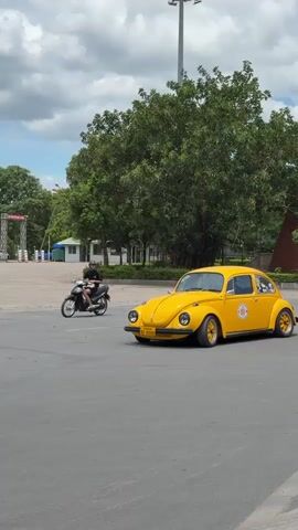 A person rides a motorcycle on a paved surface. A yellow Volkswagen Beetle is parked. A red Volkswagen Beetle with a roof rack drives past. A pink Volkswagen Beetle is parked. The background includes trees and a building. The sky is cloudy.