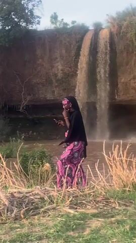 The video shows a waterfall cascading over a rocky cliff into a shallow, muddy-looking pool. A woman in a black headscarf, a patterned skirt, and black top is standing on the grassy bank near the water's edge, holding a phone. As the video progresses, the view widens to show a group of people gathered by the water, some standing and some sitting. The water in the waterfall is brown, possibly due to sediment. The surrounding environment includes green vegetation and dry, brown reeds.