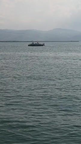 The video shows a body of water, likely a lake or sea, with gentle waves. In the distance, there are mountains and a hazy sky. A small boat with people on board is visible in the frame, moving across the water. Another sailboat is also visible further in the distance. The sun is shining, creating a shimmering effect on the water. The overall scene is serene and peaceful.