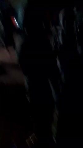 The video depicts a dimly lit outdoor scene at night. There are several people present, mostly obscured by darkness, but some are seen wearing hats or patterned clothing. Some individuals are walking and interacting with others. In the background, a building with a few visible windows is present. The video footage is somewhat shaky and lacks clarity.