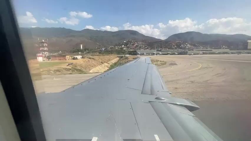The video is taken from inside an airplane, looking out the window at the wing and the airport. The plane appears to be stationary on the tarmac. The wing is grey and has many small rivets. Beyond the wing, there is a grassy area, a ditch, and an airport runway. In the background, there are buildings, hills, and a blue sky with some clouds. The video is shot on a bright, sunny day.