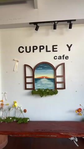 The video captures the welcoming exterior of "CUPPLE Y cafe," located in Muang Pattaya, Thailand. The scene features a light-colored wall adorned with the cafe's name in large black letters. Below the sign, a decorative wooden window frame displays a painted sunset ocean view, accented with artificial green moss and small white flowers. A rustic wooden bench sits below this decoration, also featuring moss and scattered flowers in glass vases.

To the left, the cafe entrance is a wooden door with a glass upper panel. The glass panel features a white decal reading "Welcome to CUPPLE Y," with two cartoon coffee cups forming part of the logo, and states the opening hours: "09:00 - 01:00." Adjacent to the door, a large white teacup-shaped planter overflows with colorful artificial flowers, including pink, red, yellow, and white blooms. Below this, a row of artificial red roses with green foliage rests on a white platform. White lace curtains are visible through a window next to the door. Black track lighting fixtures are mounted on the ceiling above the cafe sign, though the setting appears to be daytime. No people are visible in the video, which pans across the facade, showcasing the charming and thoughtfully decorated entrance.