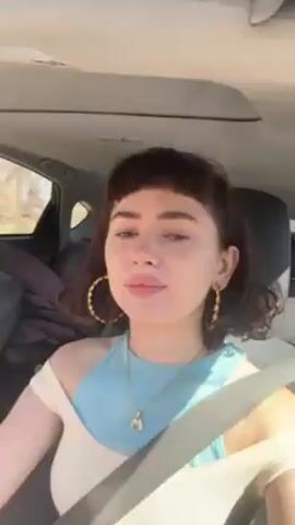 A woman with short brown hair and bangs, wearing a blue and cream top and gold hoop earrings, is driving a car. She is wearing a seatbelt and singing along to music. The background visible through the car window is a sunny, possibly rural area. There is a blanket or bag in the back seat.