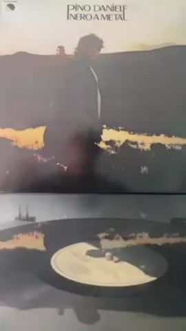 The video shows an album cover on top of a record player with the vinyl spinning. The album cover is for “Nero a meta” by Pino Daniele. The cover has an image of the musician standing in a field with a guitar on his back, staring off into the distance. The record on the player is black with a white label. The background is out of focus.