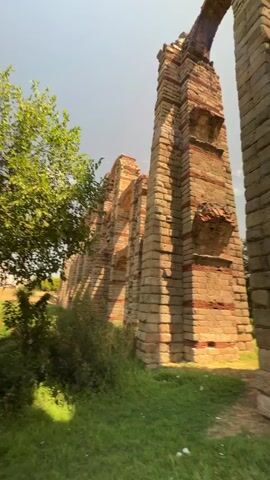 The video pans up a section of ancient Roman aqueduct, highlighting its massive stone and brick construction. Sunlight streams through the arched openings of the structure, creating dramatic shadows. Lush green grass covers the ground around the base of the aqueduct, with trees visible in the background. The camera angle is low, emphasizing the height and grandeur of the aqueduct. The sky is clear with a few wispy clouds. In the distance, two figures are visible sitting near the aqueduct.