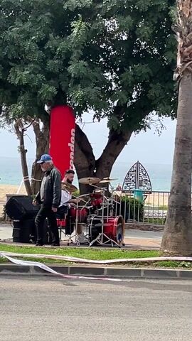The video shows a street scene by the ocean. A drummer is performing on a red drum set next to a sidewalk. A man and a woman in green jackets walk past, holding hands. A red banner stands behind the drummer. Palm trees line the street along with other greenery. There is a glimpse of the ocean in the background, and the sky is overcast. A car drives by with a person visible inside.