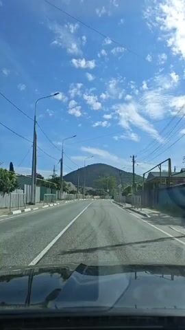 The view from inside a car shows a road heading towards a hill. The sky is blue with some clouds. There are streetlights and power lines along the road. Houses are on either side of the street.