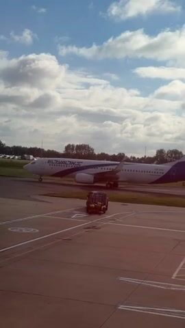 A white airplane with blue accents and the El Al logo is parked on an airport tarmac. A small white truck with a trailer is parked in front of the airplane. The sky is blue with scattered white clouds.