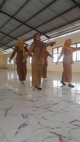Three women in matching tan uniforms and headscarves are dancing in a large, open room with a tiled floor and a corrugated metal ceiling. The woman in the center is slightly taller and has glasses. The woman on the left is shorter and is wearing white shoes. The woman on the right is also wearing dark shoes. All three women are moving their arms in a synchronized manner. The room appears to be a multi-purpose space, possibly a gymnasium or community hall, with windows visible in the background.