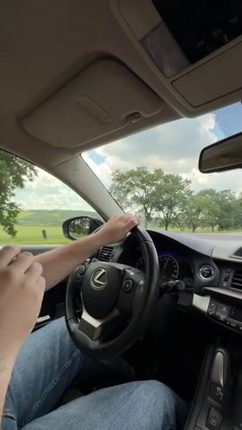The video shows a person's hands on the steering wheel of a car. The driver is wearing blue jeans and appears to be holding a phone in their left hand. The car interior is visible, including the dashboard, steering wheel with a Lexus logo, and the center console. Outside the car, trees and a cloudy sky are visible through the windows. The video appears to be shot from the passenger seat, looking towards the driver.