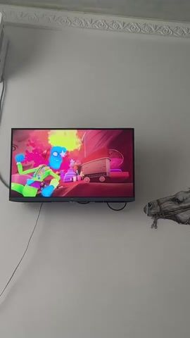 The video shows a television screen displaying an animated scene. The animation features stylized, colorful characters. In the first few seconds, a neon green and pink character with a large head is visible. The scene appears to be an interior, possibly a room with red walls and shelves. Later scenes show two characters, one black and one multi-colored, interacting. The black character resembles a cat in attire. The multi-colored character is slender with bright, flowing hair and appears to be dancing or performing. The final scene focuses on the black cat-like character, which is shown with a startled or surprised expression, wearing a white hat and suit. The overall animation style is vibrant and cartoonish, with a bright and energetic color palette. The television is mounted on a light-colored wall, and a small decorative horse head is visible on the wall to the right of the TV. A white air conditioning unit is mounted on the wall above the TV, and a black cable runs down the wall from it to the TV. The setting appears to be indoors. There is no visible text in the video. The emotions conveyed by the characters range from playful to surprised.