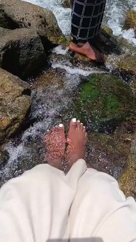 The video shows two people standing in a shallow, rocky river. The camera focuses on the feet of one person, who is wearing light-colored pants and has white nail polish on their toes. Water flows around their feet and the rocks. A second person stands behind them, wearing black and white checkered pants. Their bare feet are also in the water, positioned slightly higher up on the rocks. The environment consists of various-sized rocks, with some covered in green algae or moss, and clear, flowing water.
