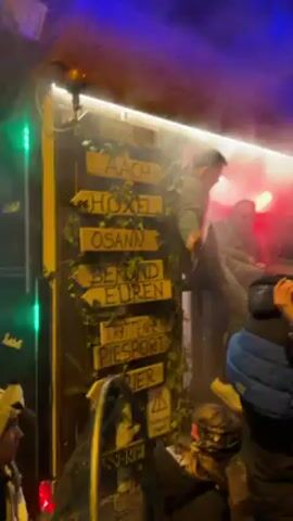 A person is standing in a booth decorated with wooden signs pointing to places, surrounded by a crowd. The booth is illuminated with white lights, and there is red and blue light further away, with fog filling the air. A speaker is visible on the side of the booth. People are watching the person in the booth. Some are holding their phones up, and the overall atmosphere is festive and crowded.
