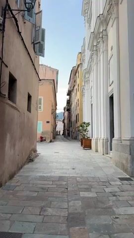 This media file captures a street scene in Bastia, France, likely during the early morning or late afternoon based on the soft lighting and shadows. The narrow cobblestone street is flanked by tall, narrow buildings with faded pastel facades. The building on the left is a light brown color, with visible electrical conduits running along its exterior. It has a single, dark-framed window. The building opposite is a vibrant peach color, with light blue shutters and a small, red fire hydrant visible at its base. Further down the street, taller buildings in shades of yellow and cream rise up, creating a sense of depth. The building on the right is a grand white structure with classical architectural elements, including columns and an arched entryway, suggesting it might be a church or a significant public building. A small, potted tree sits in a wooden planter near its entrance. The sky above is a clear, pale blue. The overall atmosphere is peaceful and quiet, conveying a sense of old-world charm. The cobblestone pavement is laid in large, irregular rectangular stones, showing signs of age and wear. There are no people clearly visible in the scene. The media appears to be a still image or a very short video clip with minimal movement.