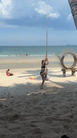 The video shows a young girl in a black bikini climbing a rope swing on a sandy beach. She is in the center of the frame, with the ocean and a cloudy sky in the background. There are other people on the beach and in the water, and a palm tree is visible on the right side of the frame. A circular wooden decoration is also visible behind the girl. The lighting suggests it is daytime, likely afternoon given the angle of the sun and the shadows. The overall mood appears relaxed and playful, with the girl enjoying herself on the swing. The scene is set in a tropical or subtropical location, indicated by the palm tree and beach setting.