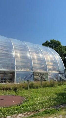 The video displays a greenhouse with a translucent, arched roof. It is a sunny day with a clear blue sky. In front of the greenhouse, there are several solar panels mounted on poles. The ground is covered with grass and some patchy vegetation. To the left of the greenhouse, there is a circular metal object. As the video progresses, the camera pans to the right, revealing more of the greenhouse's structure and the surrounding environment. Near the entrance of the greenhouse, there are some potted plants and trash cans. Further to the right, there is a small patch of trees. In the foreground, there are children's toys, including a red toy car, a yellow toy car, and a red and blue toy. There is also a black toy bulldozer. A pathway made of loose gravel leads towards the greenhouse entrance. The overall scene suggests an outdoor setting, possibly a farm or a research facility, with a focus on sustainable energy and agriculture.