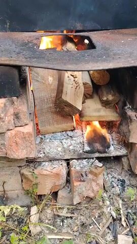 A close-up shot captures a fire burning within a makeshift brick oven. Several pieces of wood are stacked inside, with flames licking at them, producing a warm, orange glow. The fire is contained within a metal structure, possibly a large pot or cauldron, that rests on top of the bricks. The surrounding area appears to be outdoors, with some dry grass and twigs visible at the bottom of the frame. The scene is illuminated by the fire's light, suggesting it might be evening or a dimly lit area. The overall impression is one of rustic simplicity and a traditional method of cooking or heating. There is no visible text in the media file.
