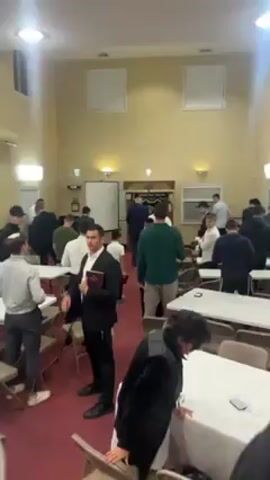 The scene is a brightly lit, moderately-sized room with beige walls and a red carpet. There are several rectangular tables covered with white cloths, surrounded by chairs. Approximately 20 to 30 people, mostly men, are scattered throughout the room. Some are standing, while others are seated. Most of the men are dressed in casual attire, although a few are wearing suits or dark jackets. One man in particular is wearing a black suit and holding a book; he gestures with his right hand as if addressing the room. A woman in a black jacket stands near the bottom right corner of the frame. The overall atmosphere seems to be one of activity and conversation, possibly before or after a meeting or event.