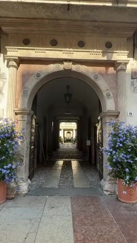 This media file captures a picturesque entryway leading to a sunlit courtyard in Trento, Italy. The scene is viewed from within a covered archway, framed by classical columns and adorned with ornate architectural details.

The subjects are primarily the setting itself. The archway, constructed from stone with decorative elements, forms a grand entrance. The pathway through the archway is paved with a mosaic of pebbles and larger rectangular stones, with patches of sunlight casting distinct shadows.

Beyond the archway, a courtyard unfolds. This space is characterized by a central fountain featuring a classical statue, flanked by potted green plants and further architectural columns. The courtyard is paved similarly to the pathway, suggesting a cohesive design.

The activity depicted is one of peaceful stillness, a moment frozen in time. The lighting suggests it is daytime, likely late morning or early afternoon, given the strong sunlight and shadows. The weather appears to be clear and sunny. The location cues, "Trento, Italy," provide specific geographical context, and the overall atmosphere evokes a sense of historical charm and serene beauty.

Notable details include the presence of a dark, wrought-iron lantern hanging from the archway's ceiling, and two large potted plants with vibrant blue flowers flanking the entrance. There is a sign with the number "19" visible on the right side of the entryway. The architectural style suggests a historic building, possibly a palace or a significant public space. The emotions evoked are those of tranquility, curiosity, and appreciation for classical aesthetics.
