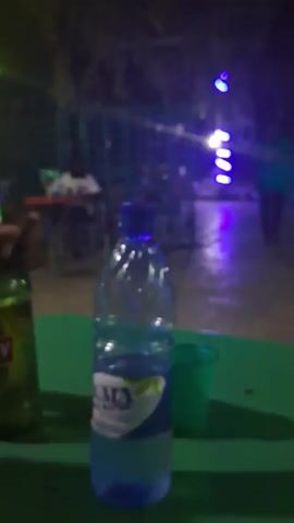 The video shows a night time scene at an outdoor bar or hangout spot. There are several people seated at tables with colorful chairs. Green tables are visible in the foreground, one with a bottle of water, a green cup, and a partially visible green beer bottle. A person's arm holding a green beer bottle is visible as the camera moves around. The area is lit with a mix of colored neon lights, including blue, yellow, and green. A fence is partially visible in the background. Overall, the setting appears to be a relaxed and social atmosphere.