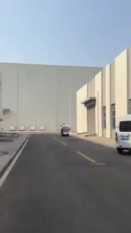 The scene shows a golf cart driving down a road with a person waving from inside it. The background shows an industrial area with large buildings. A white van is parked on the right side of the road. The day appears to be sunny and the sky is clear.