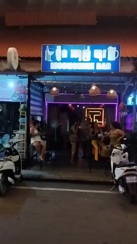 The video captures a street scene at night, likely in Southeast Asia, featuring a bar with a neon sign that reads "MOONSHINE BAR" in English, above Khmer script. The bar's entrance is illuminated with purple and white lights, and several people are visible inside, seated at tables and standing. A few motorcycles are parked on the street in front of the bar. To the right of the bar, a green food cart is visible, with an orange gas tank near it. The overall atmosphere appears lively and suggests a commercial street or entertainment district.