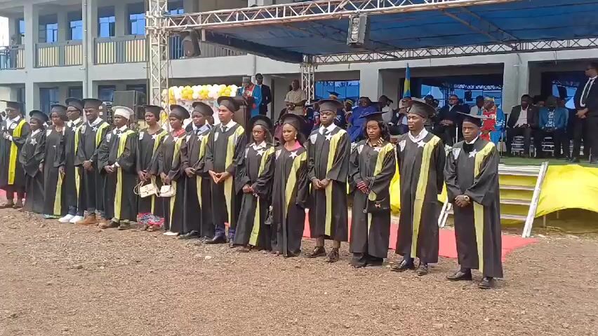 A row of about fifteen graduates stands on a gravel surface in front of a building. The graduates wear black gowns with yellow stoles and graduation caps. Behind the graduates is a raised stage with people seated on it. A red carpet leads from the graduates to the stage. The building behind the stage is a modern, two-story structure with blue window frames. The sky is clear. There are a few balloons visible in the background. A car is parked on the left side of the frame.