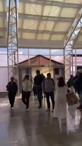 The video shows a group of people walking through what appears to be an airport or a large indoor waiting area with high ceilings supported by metal framework. Sunlight filters through the ceiling's translucent panels.  Most people walk towards an exit at the end of the hall.  The floor is tiled in a gray, marbled pattern. The individuals are dressed in casual clothing. One man wears a white t-shirt with the word "BOSS" printed on it and carries a neck pillow.  Several other people can be seen in the background. Some are carrying luggage or backpacks. The overall atmosphere seems calm and orderly.