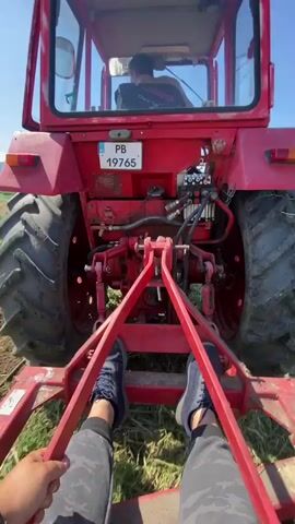 A first-person perspective video shows a red tractor with a person driving it. The camera is positioned behind the tractor, attached to a farming implement. The tractor is moving through a field of green grass and tilled soil. The driver is wearing a black shirt and dark pants. The camera person is wearing dark sneakers and patterned pants, holding onto red metal bars. The tractor has a license plate that reads "PB 19765." In the background, there are trees and a distant hillside with some structures. The sky is blue with a few white clouds. The farming implement behind the tractor appears to be a disc harrow or similar tillage tool, churning the soil and grass.