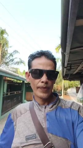 A man with short, dark hair, sunglasses, and a gray and blue jacket is standing outdoors, facing the camera. He has a brown strap across his chest. Behind him, there is a green fence, palm trees, and a building with a corrugated roof. The sky is visible above. The scene appears to be a residential or rural area.