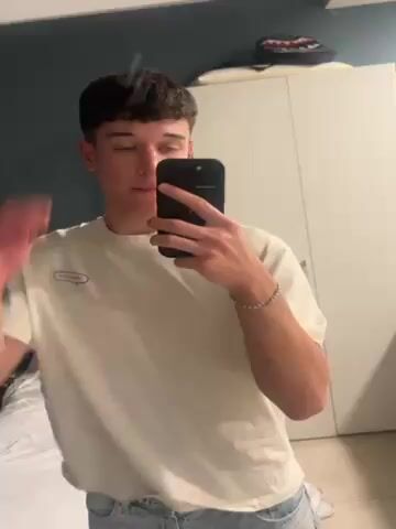 A young man is recording a selfie video in front of a mirror in what appears to be a bedroom. He is wearing a light-colored t-shirt, faded blue jeans, and white sneakers. He runs his hand through his dark, curly hair at the start of the video. The bedroom has a bed with white linens, a dark blue wall, and a white closet. The man holds a black smartphone, presumably recording the video.