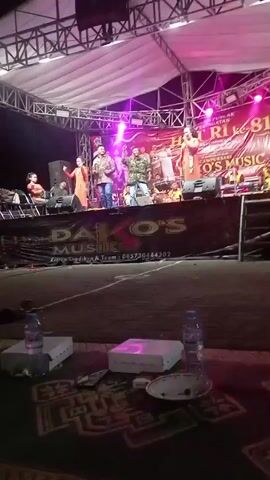 A live music performance is underway on an outdoor stage. The stage is illuminated by bright pink and blue lights. Several individuals are visible on the stage, including performers singing and playing instruments, as well as other people standing around. A large banner with text and a logo is displayed behind the performers. In the foreground, several water bottles and food containers are placed on what appears to be a patterned mat or carpet. The overall atmosphere suggests a lively outdoor event.