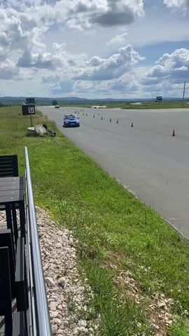 A blue Subaru Impreza WRX STI is seen driving on a paved track. The car has a large spoiler and racing decals. The track is lined with orange traffic cones and white barriers. In the background, there is a large white tent with Russian text that reads "ТРЕНИНГ-ЦЕНТР САМАРА" (TRAINING CENTER SAMARA). The sky is cloudy, and there are green grassy areas on either side of the track. The car appears to be participating in some form of automotive event, possibly a track day or autocross.
