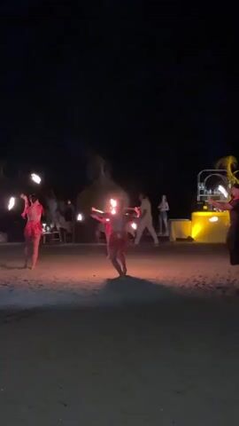 This media file captures a nighttime scene on a sandy beach, likely a resort or entertainment venue, with a performance taking place. The primary subjects are two individuals, dressed in what appear to be vibrant, sequined costumes, actively engaged in a fire-spinning or fire-dancing performance. Both are holding objects that are alight, creating dramatic trails of fire and smoke that illuminate the immediate area. One performer in the center is spinning a hoop of fire above their head, while the other on the left is twirling flaming batons.

In the background, other people are visible, some seated at tables and others standing, observing the performance. A bar area with bright yellow lighting is also present to the right, suggesting this is an evening entertainment event. The setting is a beach, indicated by the sand underfoot and the dark, open sky above. The time of day is clearly night, with artificial lights providing illumination and the performers' fire being the most prominent light source. The atmosphere appears festive and engaging, with the fire performance creating a sense of excitement and spectacle. There is no visible text in the media file.