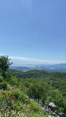 The video opens with a wide shot of a vast green landscape under a clear blue sky. Lush green hills and mountains stretch into the distance, with a body of water and more mountains visible on the far horizon. In the immediate foreground, green foliage and some rocks are present. Around the three-second mark, a person with red hair, wearing a black top, briefly enters the bottom right corner of the frame, looking towards the right side of the view before the video ends. A vehicle sound is audible throughout the clip.