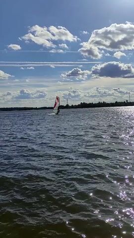 A windsurfer is on a body of water, moving from left to right across the screen. The windsurfer is standing on a board with a sail. The sail is white with red markings. The water is rippled and appears dark blue. The sky is partly cloudy with blue patches. There is a tree line in the background and a tall tower in the distance. The sun is shining and reflecting off the water. The video is shot from a low angle, close to the water.