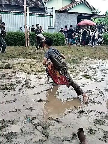 The video shows two people in a muddy field sparring. There are other people standing around watching. The spar is done with what appear to be padded cylinders, which the participants use to hit each other. At one point, one participant falls to the ground in the mud.