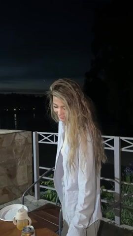 A woman with long, blonde, wavy hair is standing on a balcony at night. She is wearing a light blue unbuttoned shirt over a cream-colored top and light-colored pants. Her head is tilted down and she appears to be looking at something in front of her. There is a railing behind her and a body of water with lights visible in the distance. On a table in the foreground, there is a white plate, a sippy cup, and a can of what appears to be a beverage. The woman then looks up and smiles, extending her left arm slightly outward.