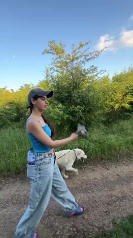 A young woman with long dark hair, a baseball cap, a blue tank top, and baggy jeans is walking a light-colored dog on a leash along a dirt path lined with green foliage. She has a water bottle in one of her back pockets and is carrying something in her hand. The sky is blue with some white clouds.