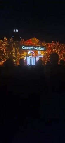 The video shows a wide shot of an outdoor nighttime event where a building is projected with light. A crowd of silhouetted figures is visible in the foreground, facing the building. The projection on the building features text that reads "MARS!" and "Mobilizing Awareness for Resilient Societies". Below this, there are dates or numbers that appear to be "15.-27.10.26" followed by "Karlsruhe". The lighting is predominantly blue and white, illuminating the building and casting a faint glow on the audience. Two lamps are visible hanging in the air above the building.