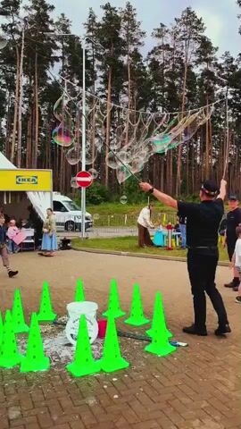 A group of people are outdoors making giant bubbles. A man in a dark shirt and pants holds two long poles with a string in between them, creating a large, colorful bubble. Several children are playing around the bubbles, trying to catch or pop them. Another adult is visible in the background, possibly assisting with the bubble-making. There are green cones and white buckets on the ground, suggesting a setup for this activity. In the distance, a forest of tall trees is visible, along with a yellow sign that appears to say "IKEA" and a white van parked on a paved road. The sky is overcast.