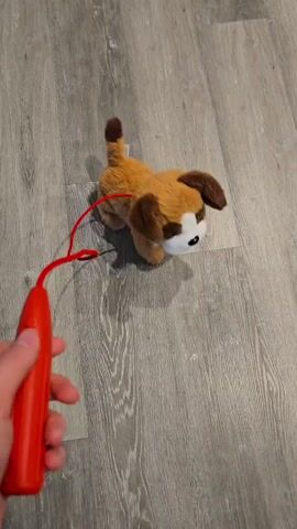 A person holds a red leash attached to a stuffed toy dog. The dog has brown and white fur. The dog is on a grey wooden floor and appears to be walking as the leash is pulled.
